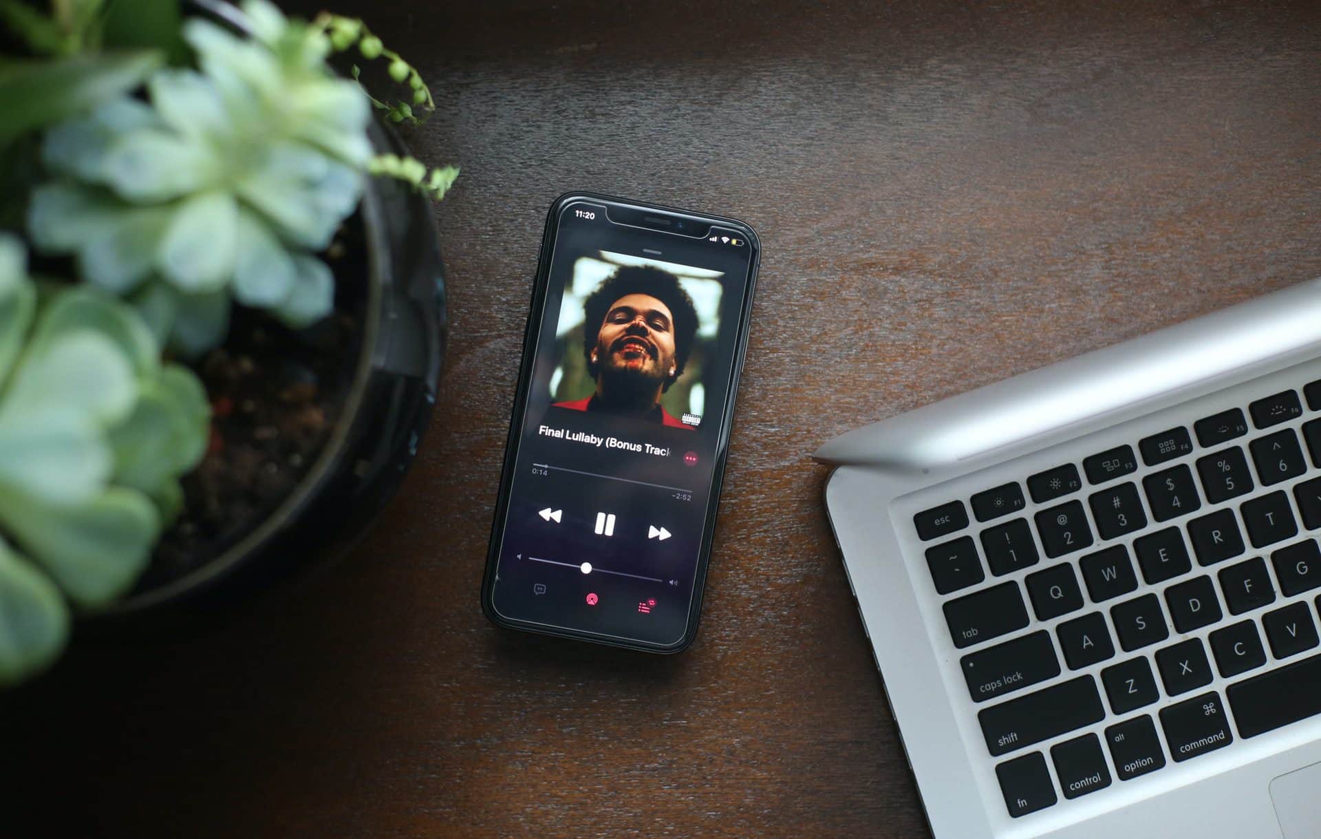 Aplikácia Apple Music na iPhone, ktorý je položený na stole vedľa kvetináča a strieborného MacBooku Air