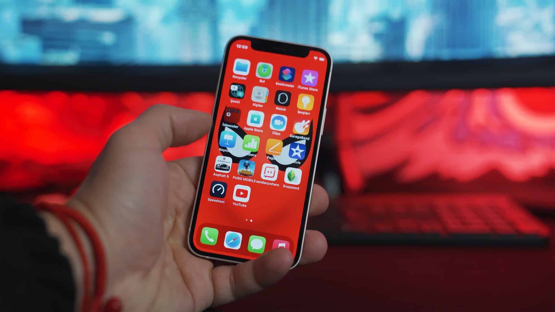 iPhone 11 s červeným pozadím a aplikáciami v ruke na červeno-modrom pozadí. Vidieť aj monitor a klávesnicu