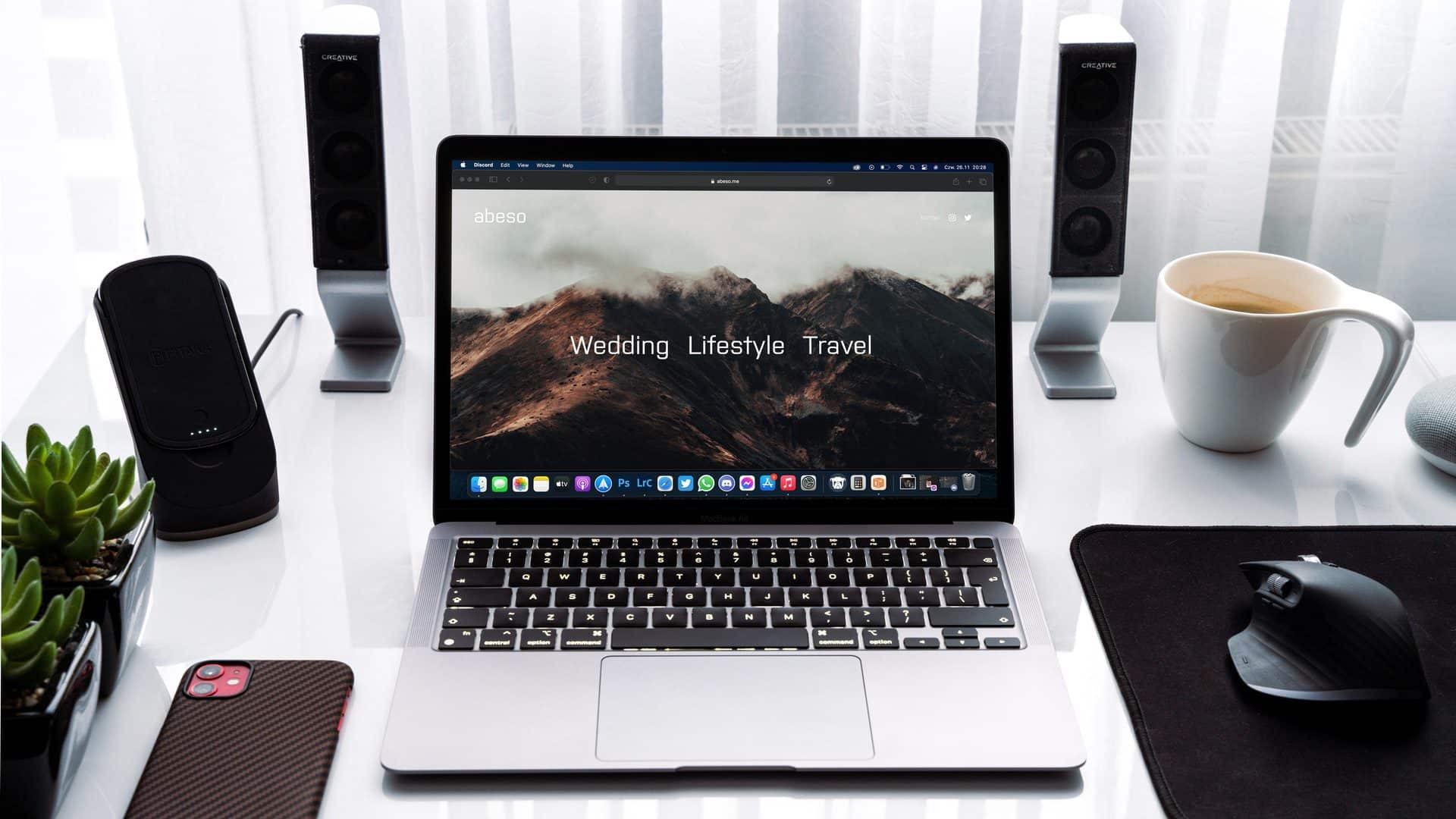 MacBook Air 2020 M1 na stole. Položený je tu aj iPhone, káva či myš s reproduktormi