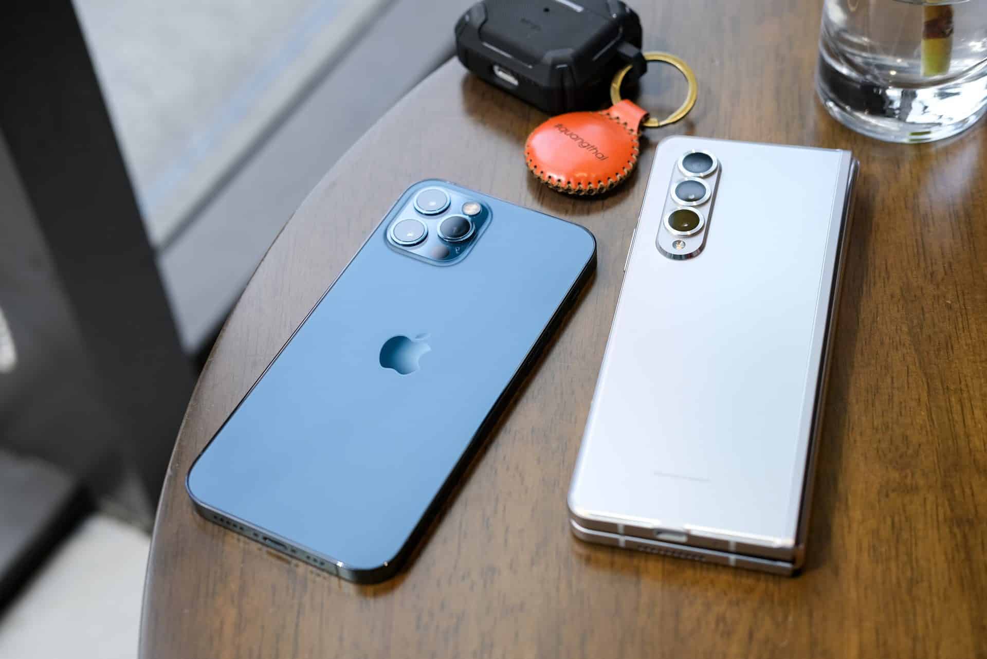 iPhone 12 Pro Max v modrej farbe a Samsung Galaxy Z Fold 3 v bielej farbe na stole vedľa AirTagu a AirPods