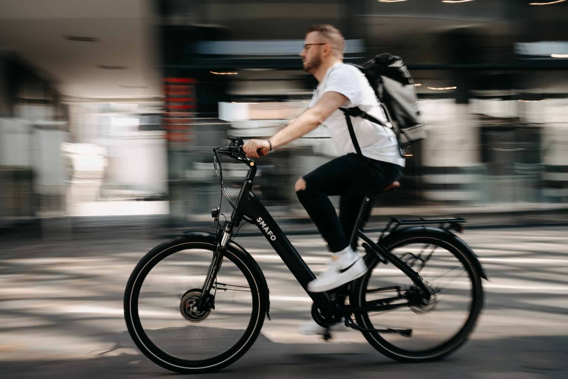 Muž v bielom tričku, s čiernymi nohavicami a bielymi teniskami Nike jazdiaci na elektrickom bicykli v meste